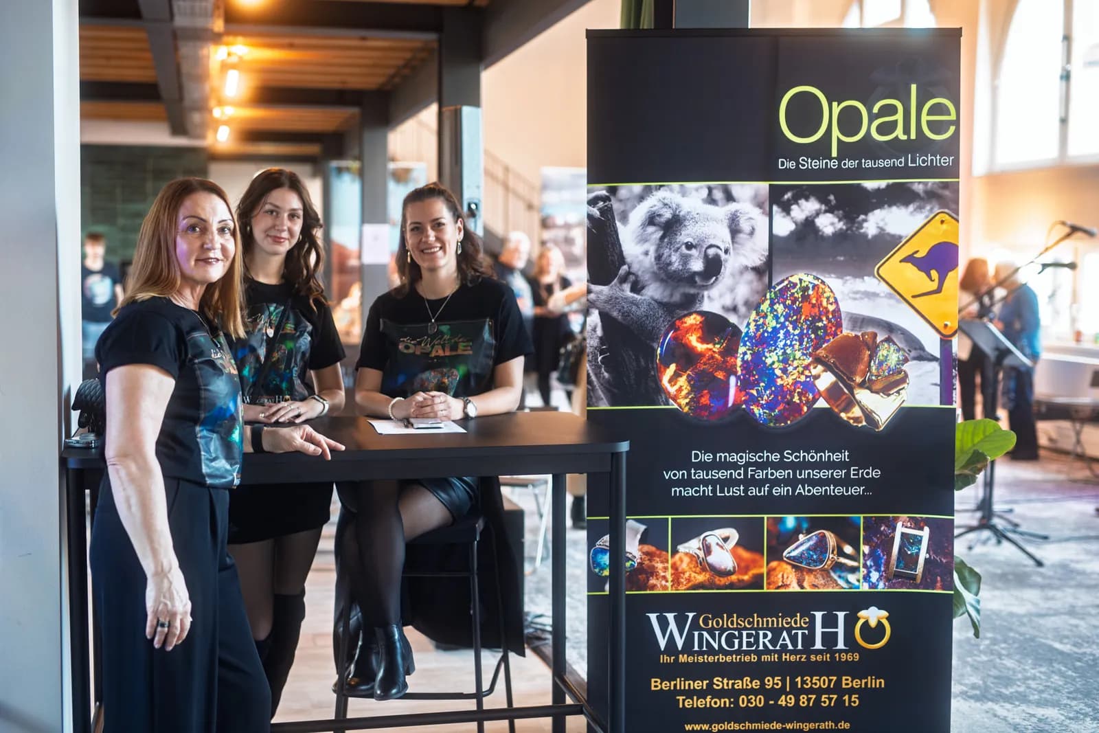 Empfang im Postwerk: drei Frauen in Opal-T-Shirts am Tisch neben dem Ausstellungsbanner